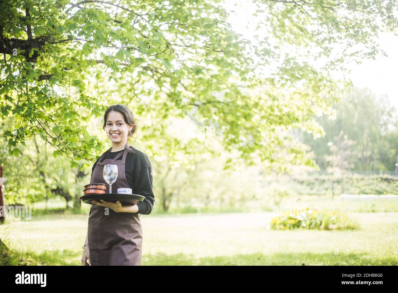 Portrait d'une serveuse jeune femme confiante souriant tout en tenant le service plateau contre les plantes Banque D'Images Portrait d'une serveuse jeune femme confiante souriant tout en tenant le service plateau contre les plantes Banque D'Images