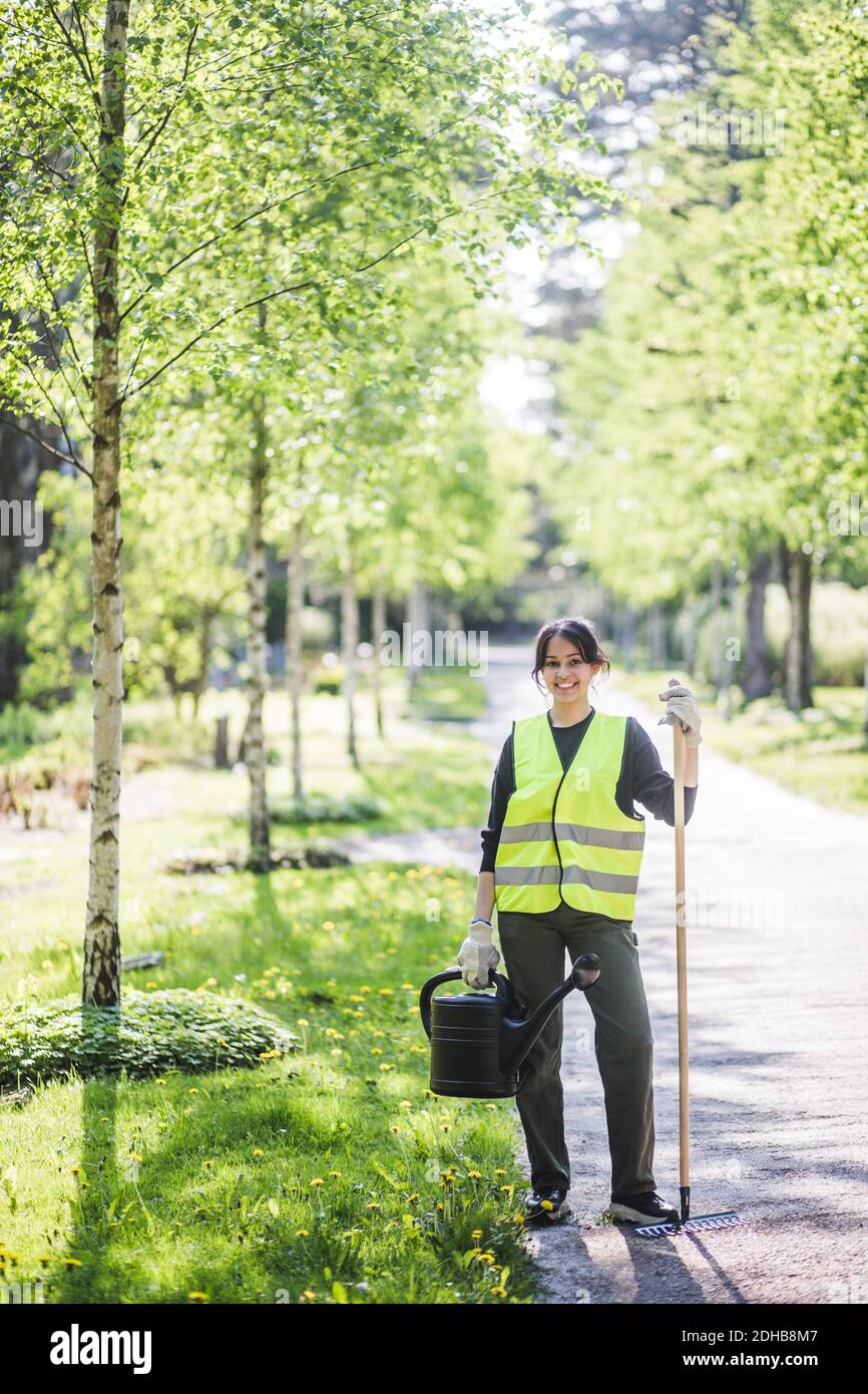 Portrait complet de jeune femme souriante tenant un arrosoir et râteau sur le sentier au jardin Banque D'Images