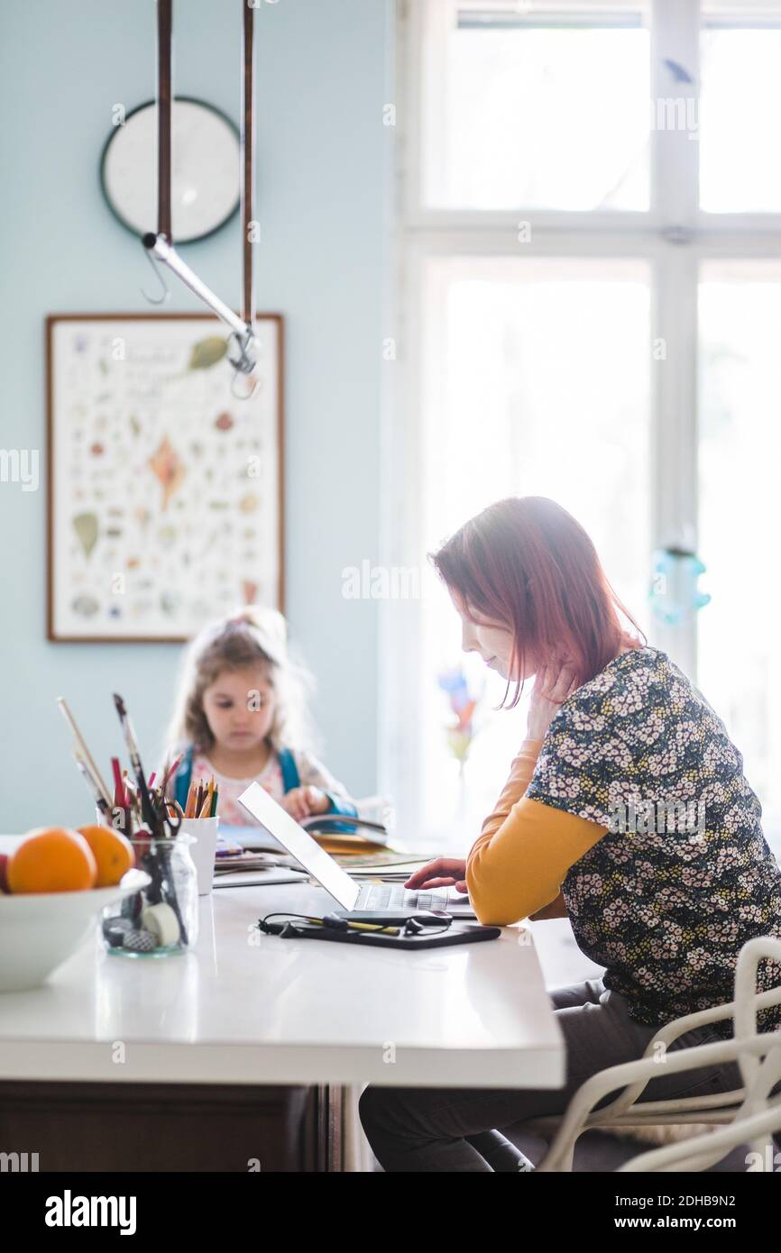 Vue latérale des femmes de taille moyenne travaillant sur un ordinateur portable assis avec une fille à kitchen island Banque D'Images