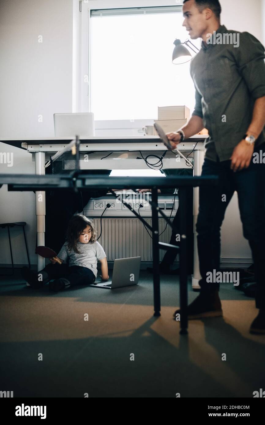 Toute la longueur d'homme d'affaires jouant au tennis de table pendant que le garçon utilise ordinateur portable au bureau créatif Banque D'Images