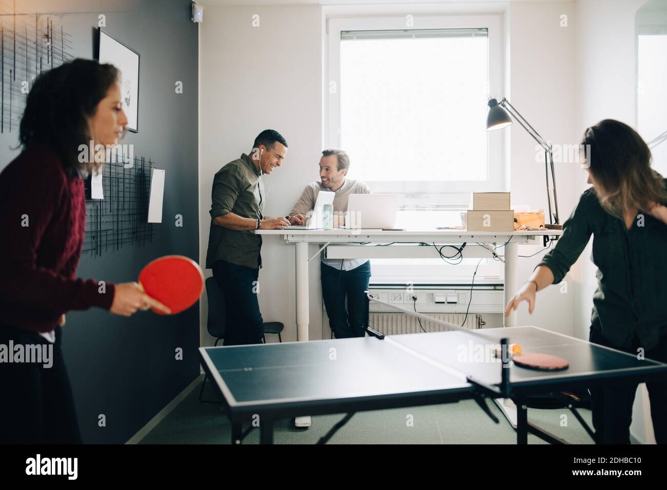 Les femmes jouent au tennis de table tandis que les collègues masculins utilisent un ordinateur portable au bureau de création Banque D'Images