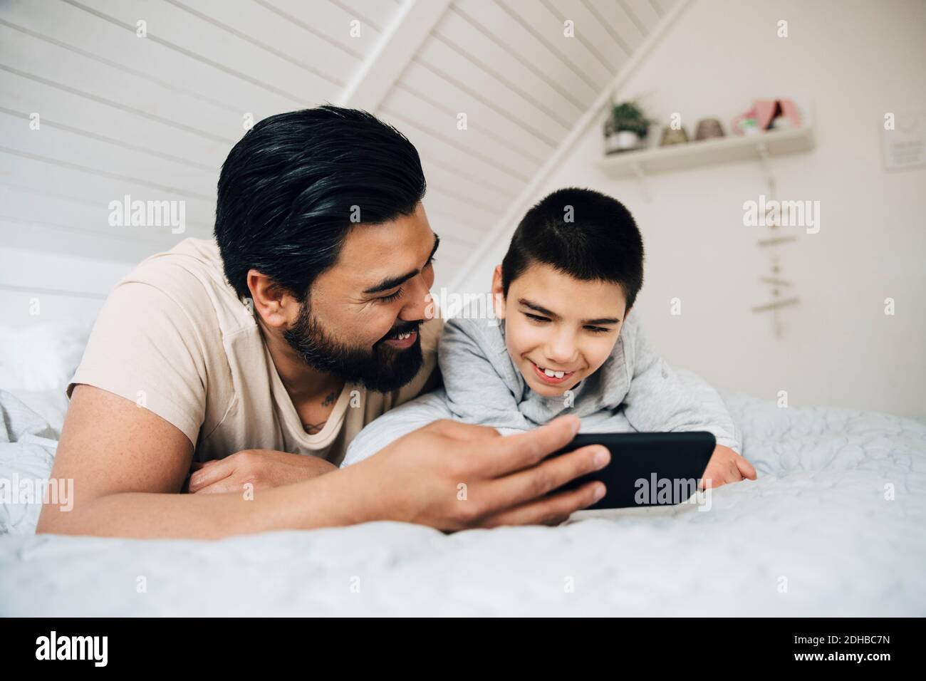 Père et fils handicapés souriants regardant un film sur un téléphone portable en étant allongé sur le lit à la maison Banque D'Images