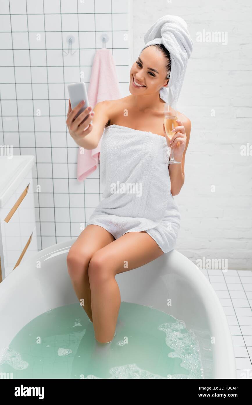 Femme souriante avec un verre de champagne prenant le selfie dans la salle de bains Banque D'Images