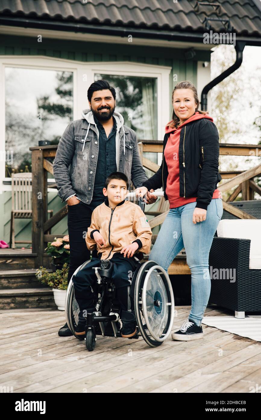 Portrait de parents souriants debout par un fils handicapé assis fauteuil roulant contre la maison Banque D'Images