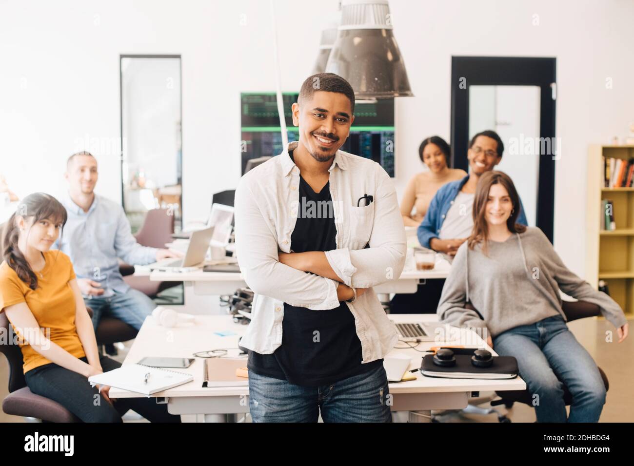 Portrait d'un pirate informatique confiant avec les bras croisés debout pendant des collègues heureux assis au bureau Banque D'Images