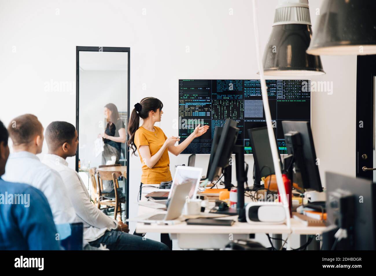 Hacker informatique féminin présentant du code sur l'écran de télévision pendant la réunion avec des programmeurs dans le bureau de création Banque D'Images