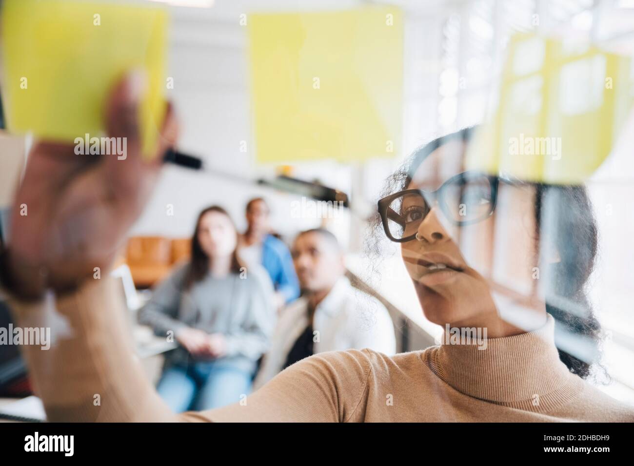 Hacker informatique femelle collant des notes adhésives sur le verre avec des collègues assis en arrière-plan Banque D'Images