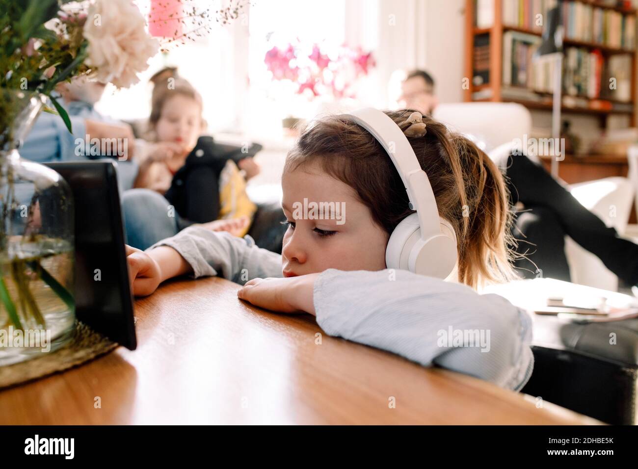 Une jeune fille écoute de la musique à l'aide d'un casque et d'une tablette numérique avec famille en arrière-plan Banque D'Images