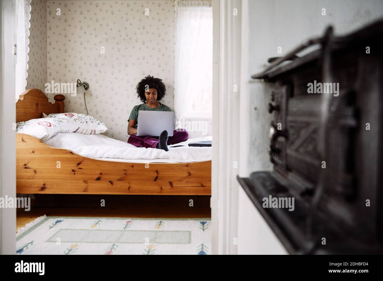 Jeune femme utilisant un ordinateur portable tout en se relaxant sur le lit à la maison vue par la porte Banque D'Images