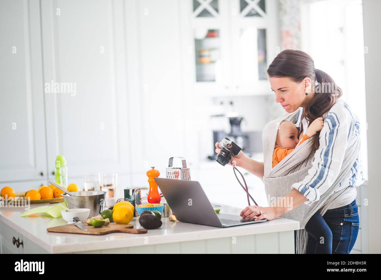 Vue latérale de la nourriture de la mère qui fonctionne blogging pendant l'allaitement de la fille dans un porte-bébé dans la cuisine Banque D'Images