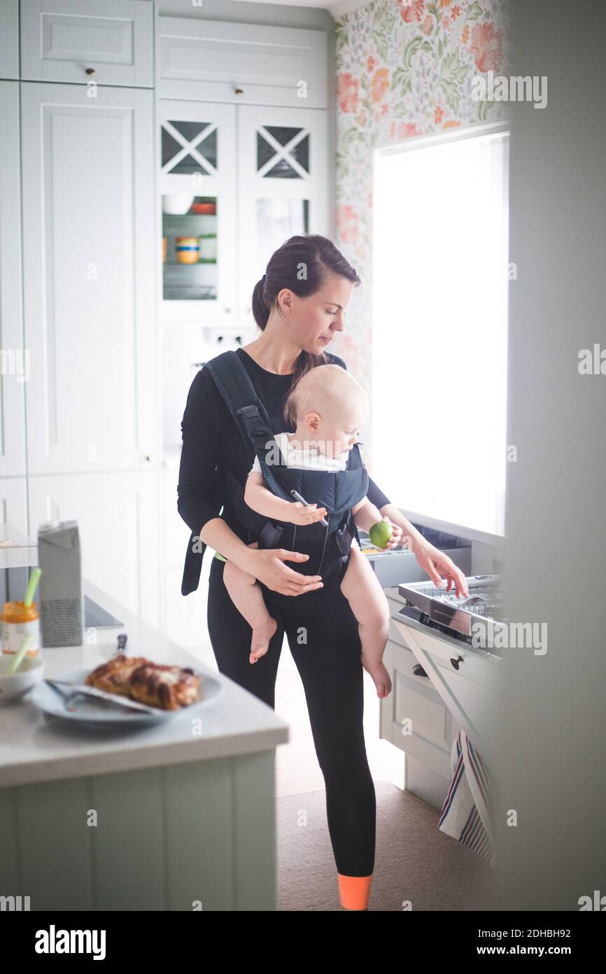 La mère porte sa fille dans un porte-bébé tout en étant debout dans la cuisine à la maison Banque D'Images