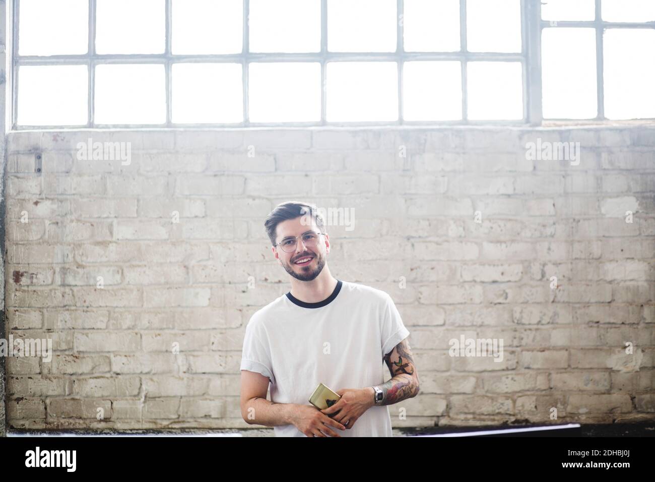 Un jeune pirate informatique souriant qui regarde loin tout en se tenant avec intelligence téléphone contre mur de briques dans le bureau Banque D'Images