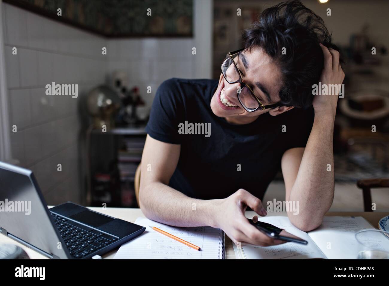 Joyeux média social accro jeune homme utilisant le téléphone mobile pendant faire des devoirs Banque D'Images