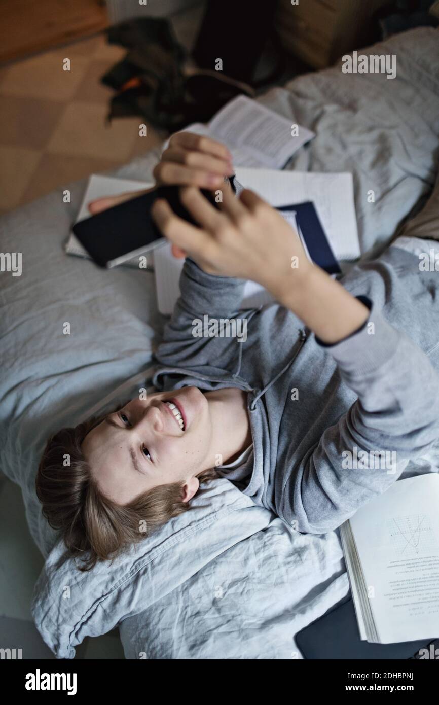 Vue en grand angle d'un adolescent souriant à l'aide d'un téléphone portable tout en étudiant dans la chambre Banque D'Images