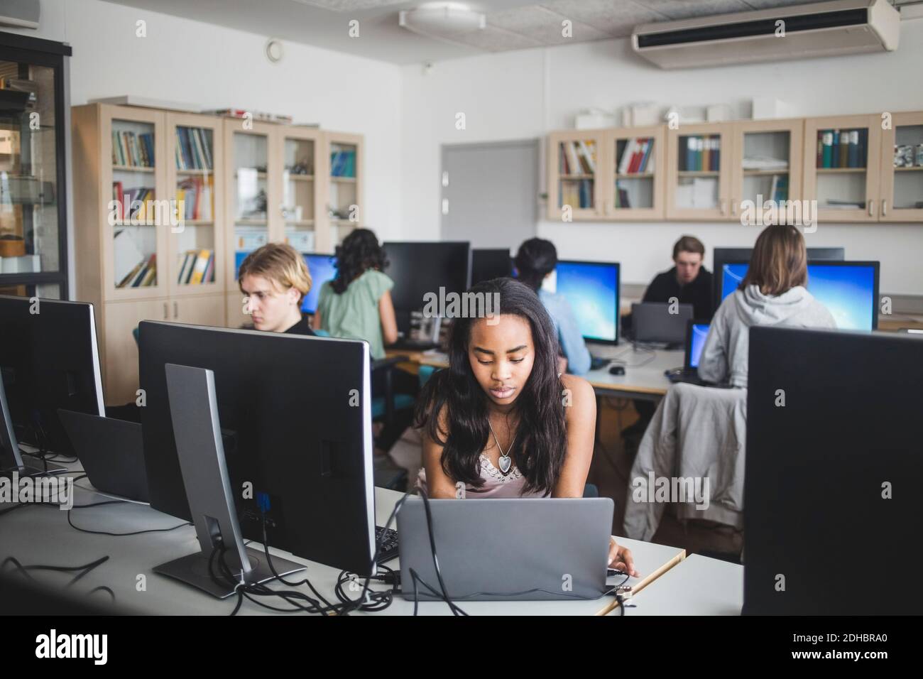 Une lycéen de sexe féminin utilisant un ordinateur portable au bureau contre un enseignant et des amis assis dans un laboratoire informatique Banque D'Images