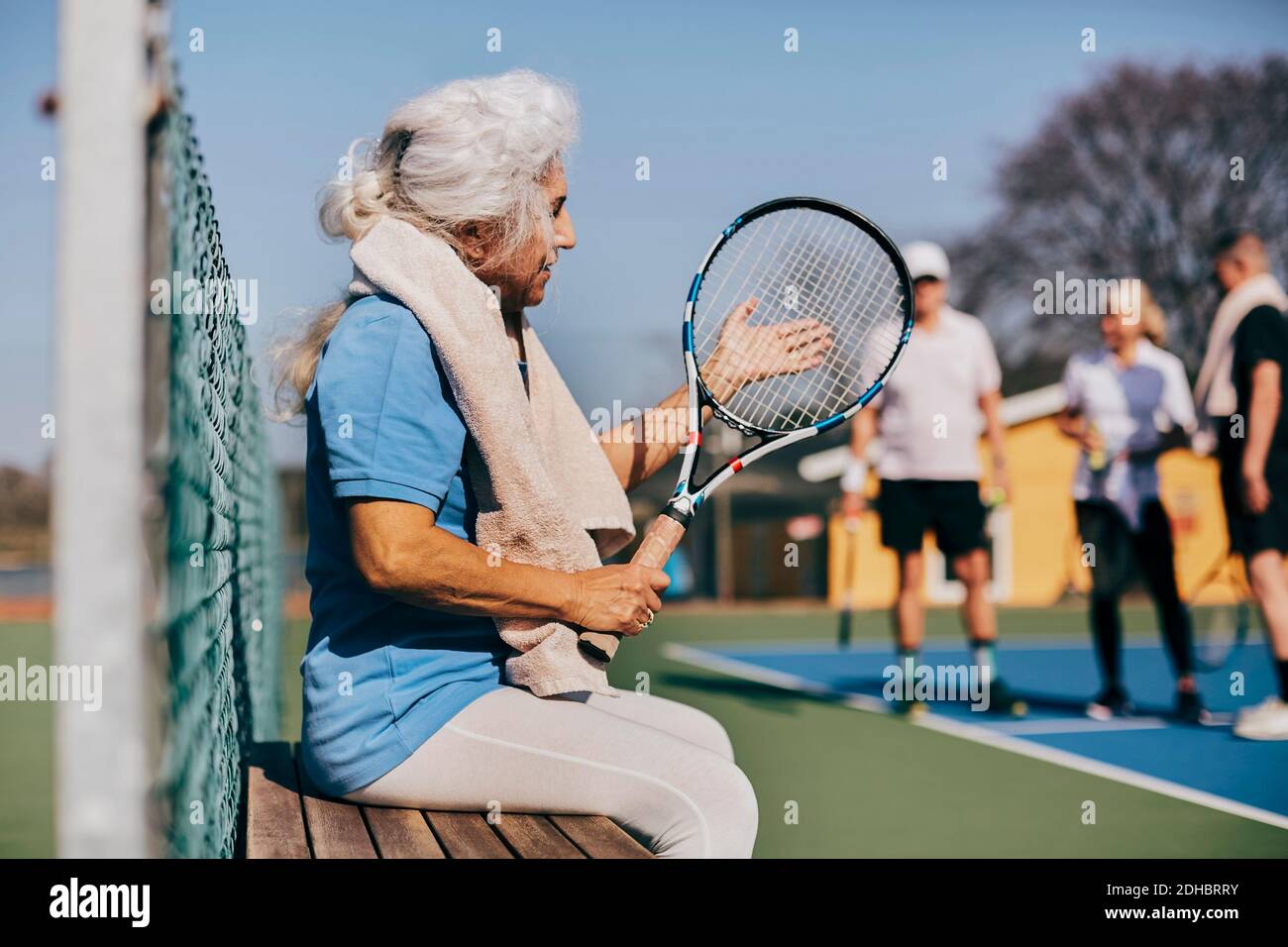 Femme âgée tenant une raquette de tennis tout en étant assise sur le banc à court de tennis Banque D'Images