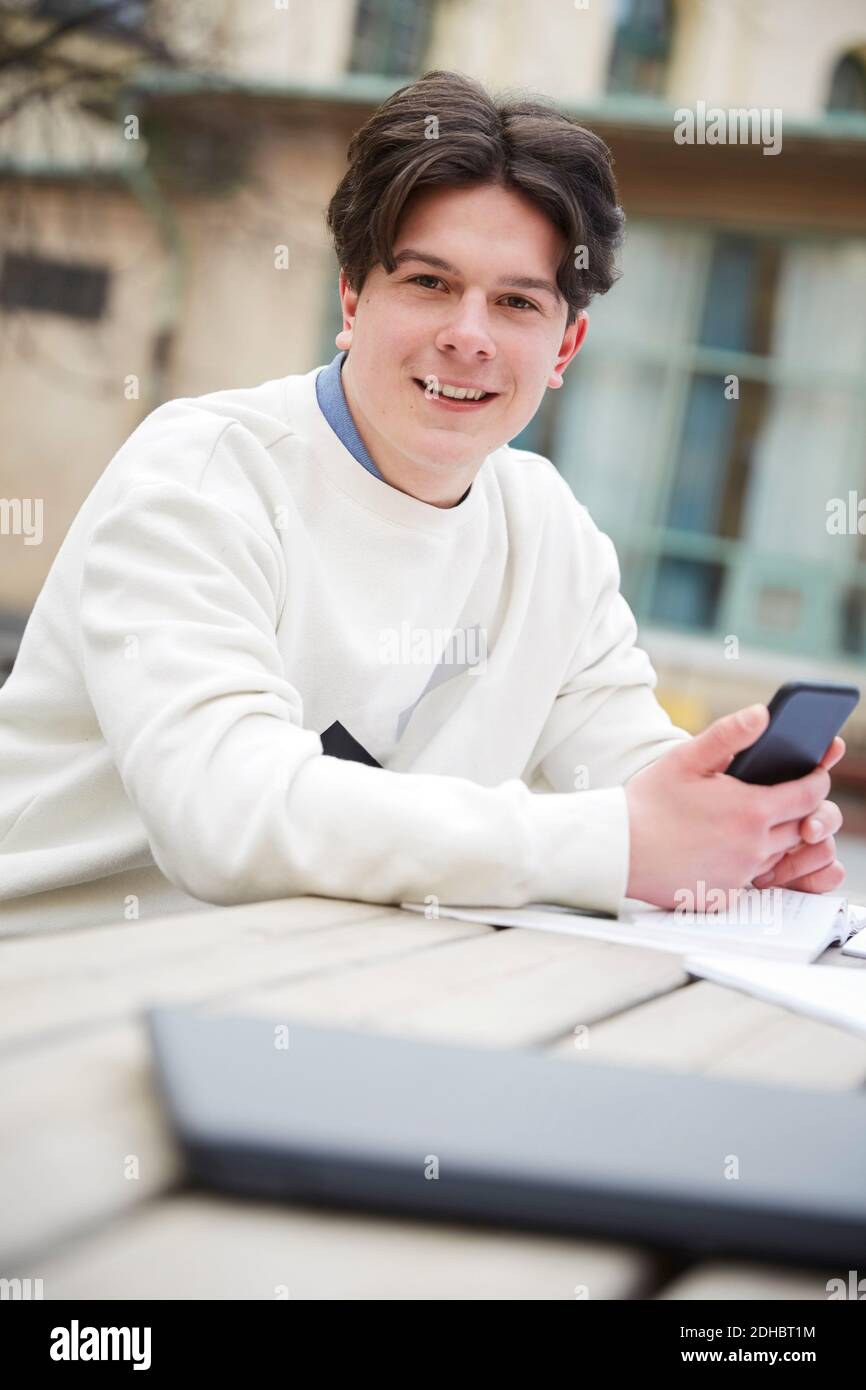 Portrait d'un adolescent confiant tenant un téléphone portable assis à la table dans la cour d'école Banque D'Images