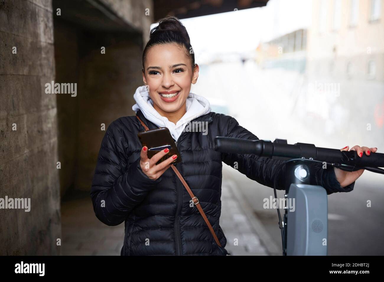Adolescente souriante avec scooter électrique poussant en regardant loin tenir le téléphone mobile sur la voie de passage Banque D'Images