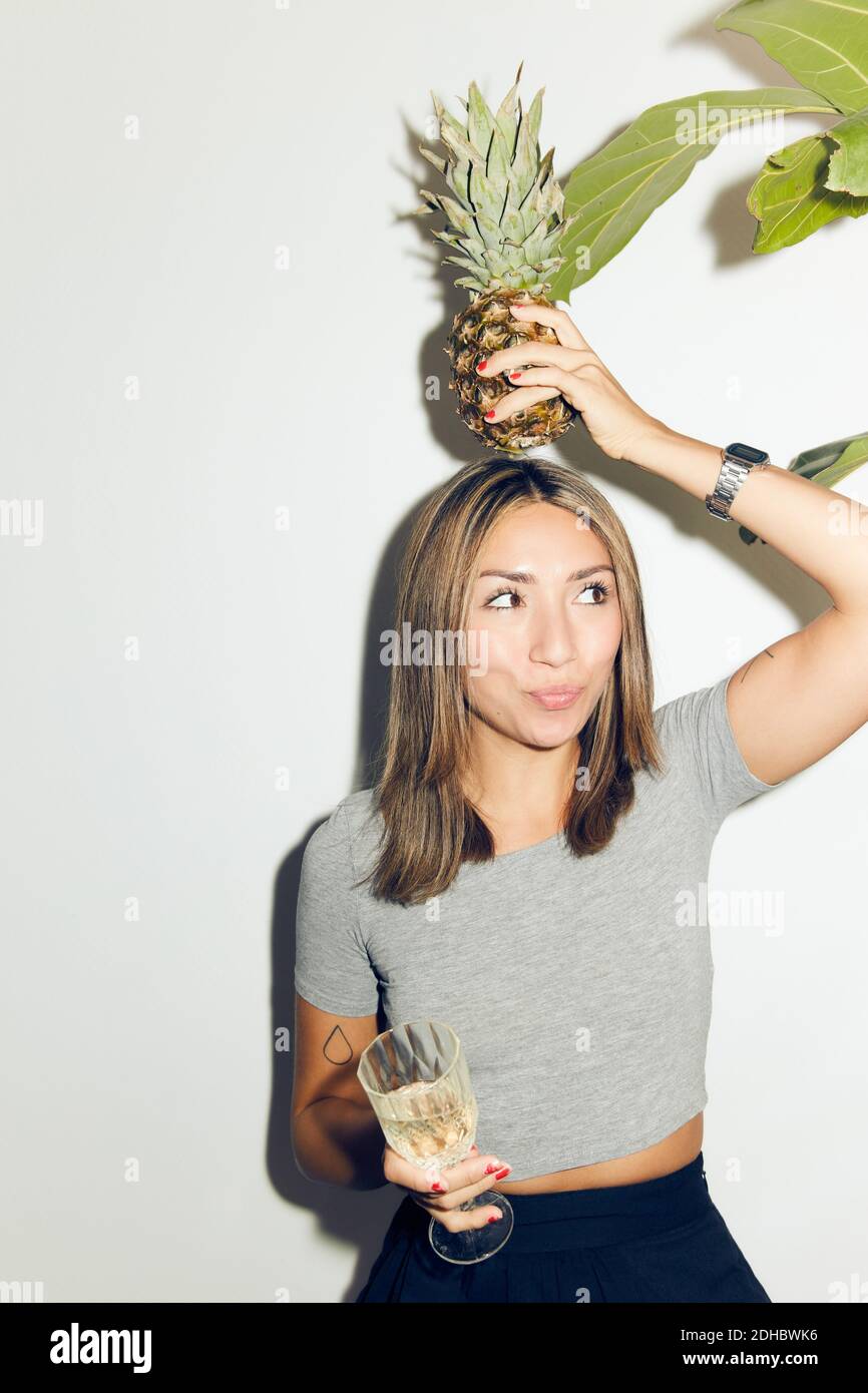 Jeune femme joueur portant de l'ananas sur la tête tout en étant debout avec buvez contre le mur blanc à la maison Banque D'Images