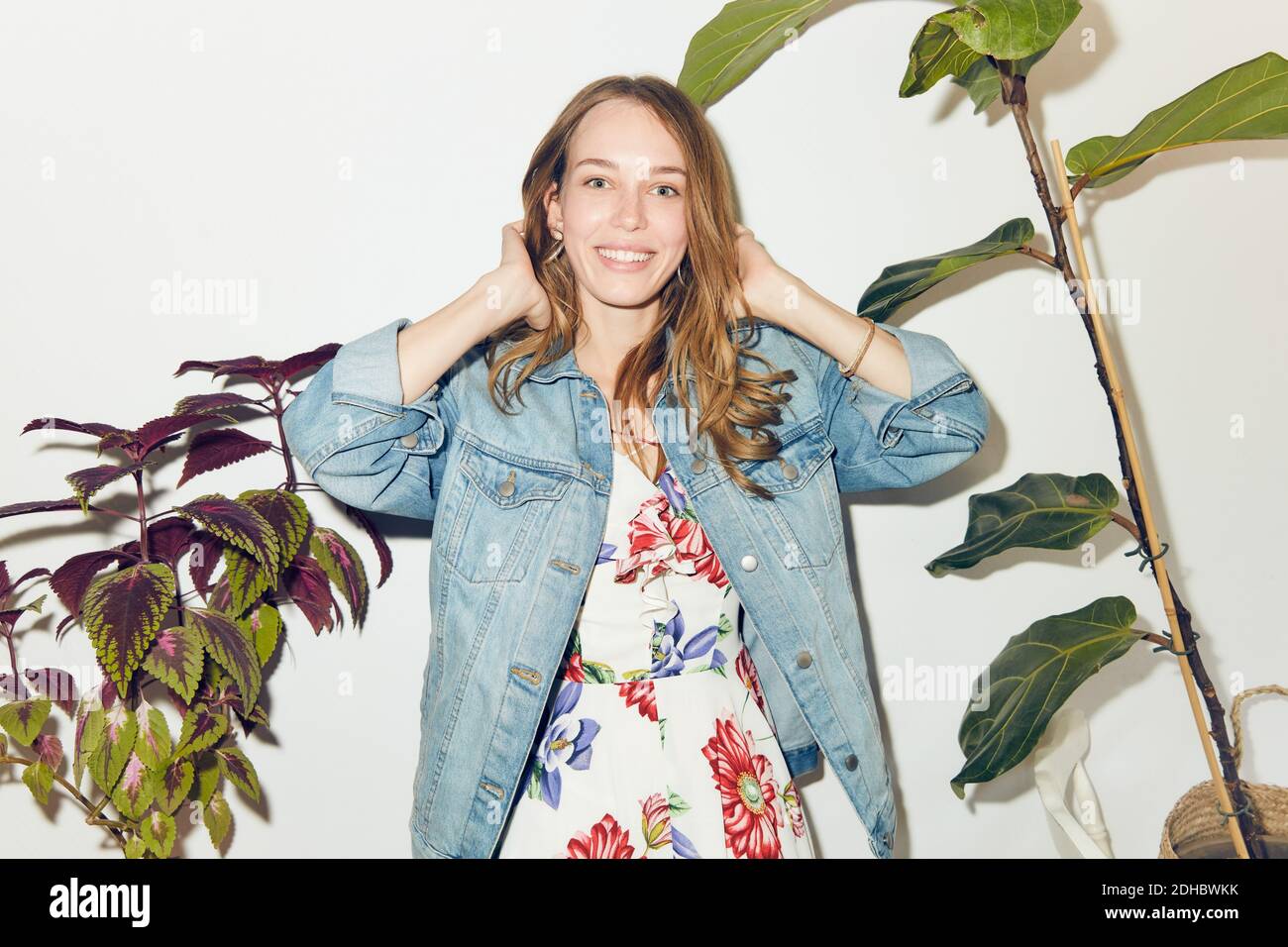 Portrait d'une jeune femme souriante avec les mains dans les cheveux debout au milieu des plantes contre le mur blanc à la maison Banque D'Images