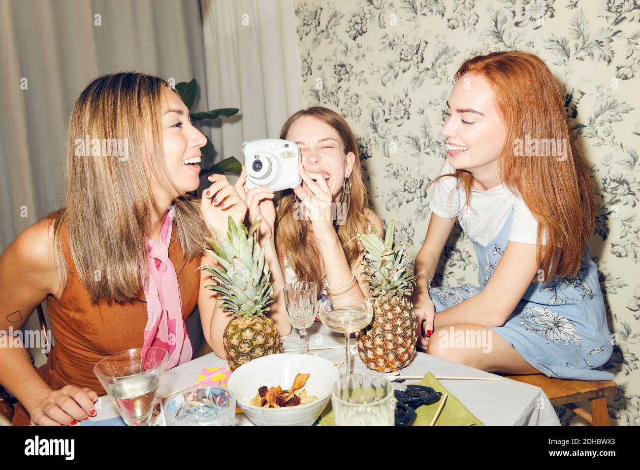 Jeune femme joyeuse tenant un appareil photo tout en étant assise parmi des amies féminines à la maison pendant le dîner Banque D'Images