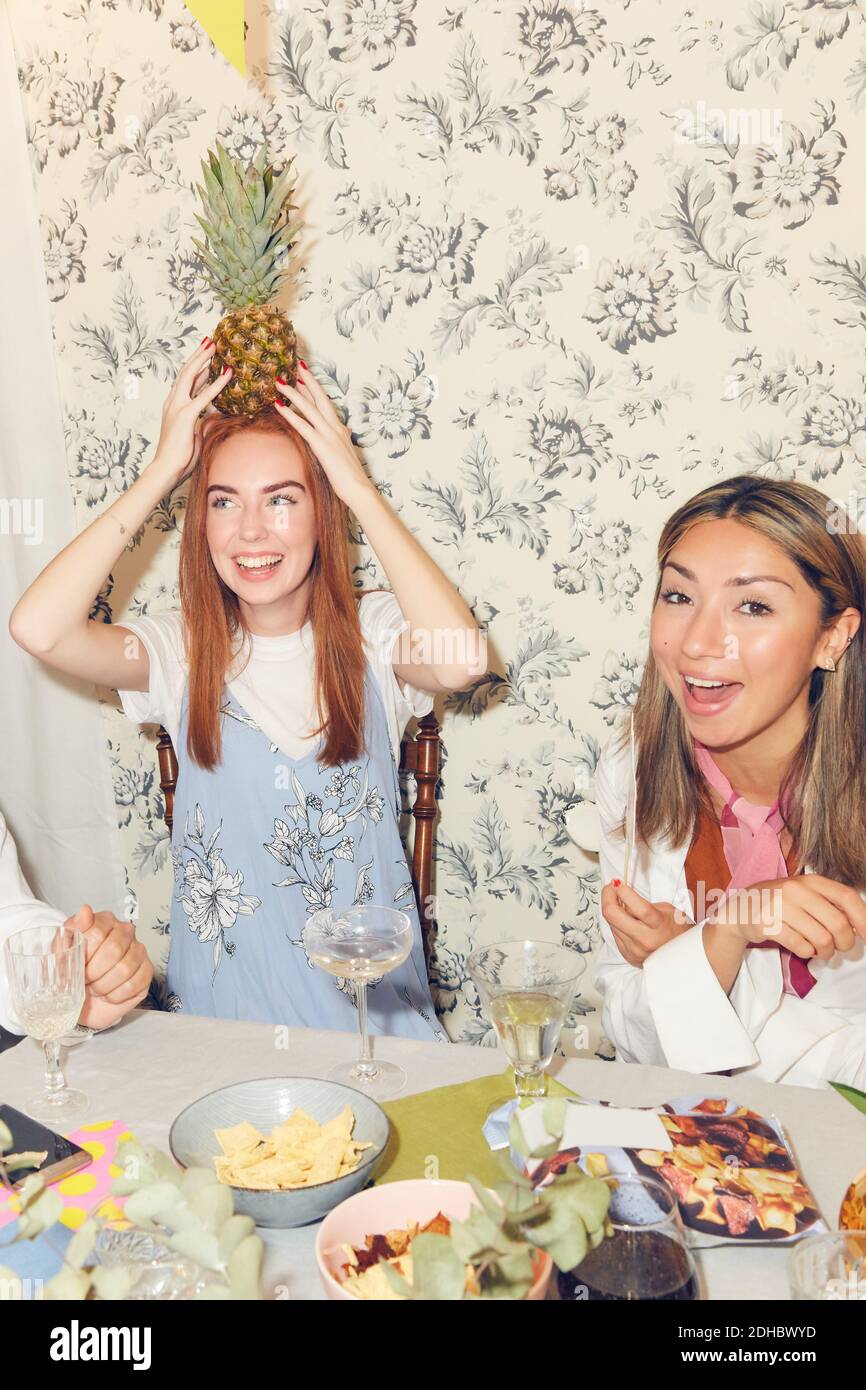 Jeune femme joueur tenant l'ananas sur la tête tout en étant assise avec amis à la table à manger pendant la fête dans l'appartement Banque D'Images