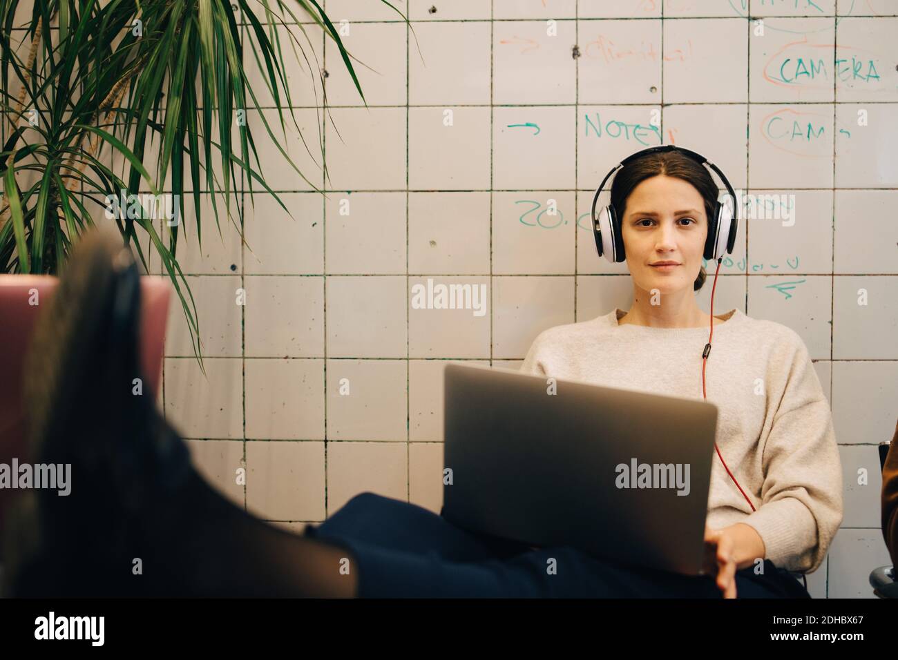 Portrait d'une jeune femme confiante qui écoute des hacker informatiques à l'aide d'un casque tout en étant assis avec un ordinateur portable contre le carrelage w Banque D'Images