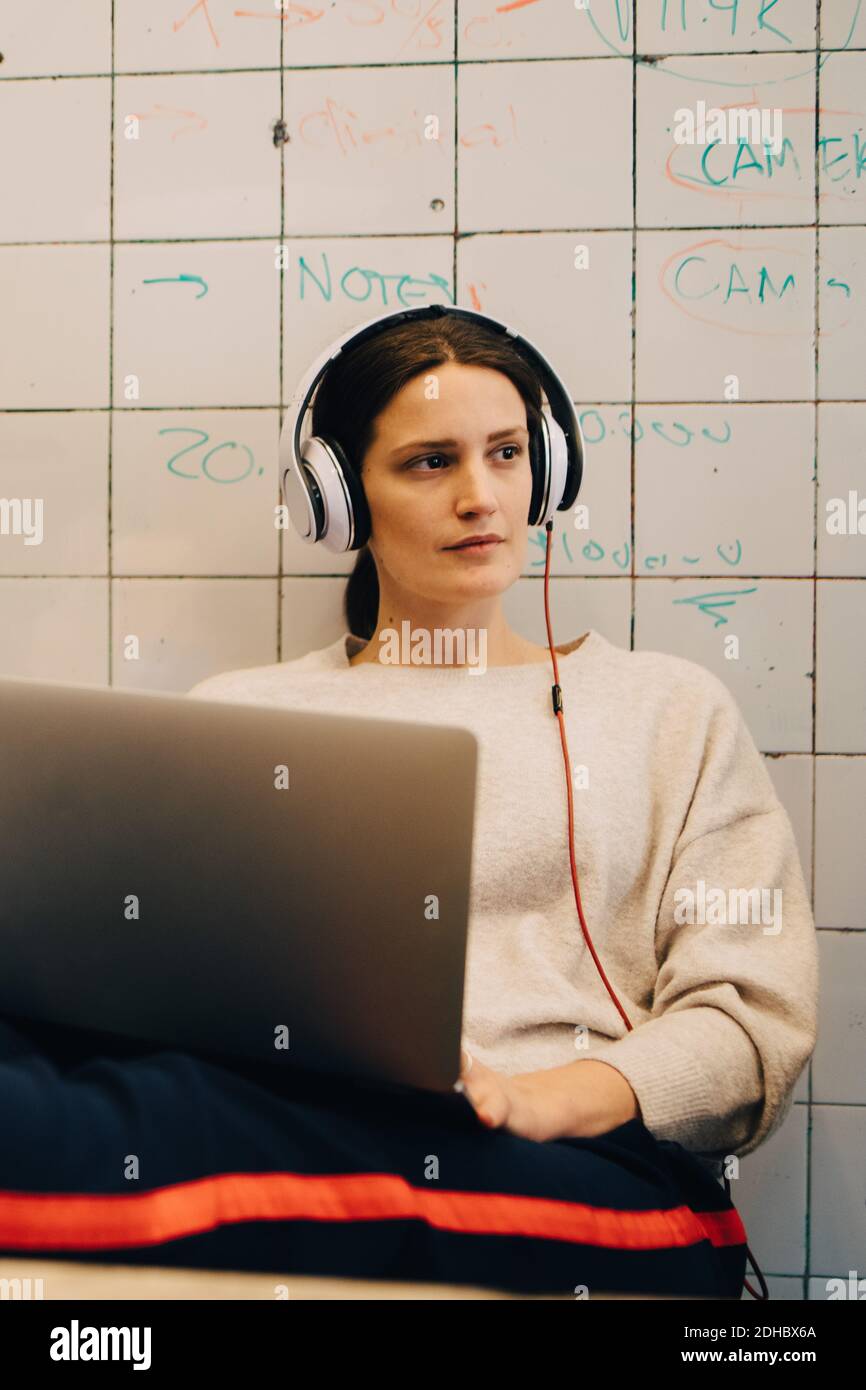 Jeune femme hacker d'ordinateur à l'écoute par le casque pendant qu'elle est assise avec ordinateur portable contre un mur de carreaux dans un bureau créatif Banque D'Images
