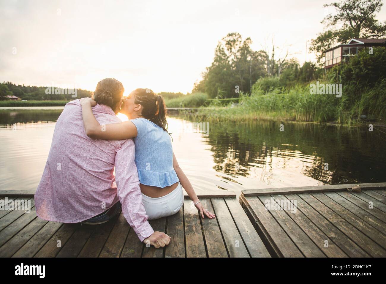 Vue arrière de l'ami embrassant petite amie assis avec les bras autour sur la jetée au-dessus du lac pendant le coucher du soleil Banque D'Images