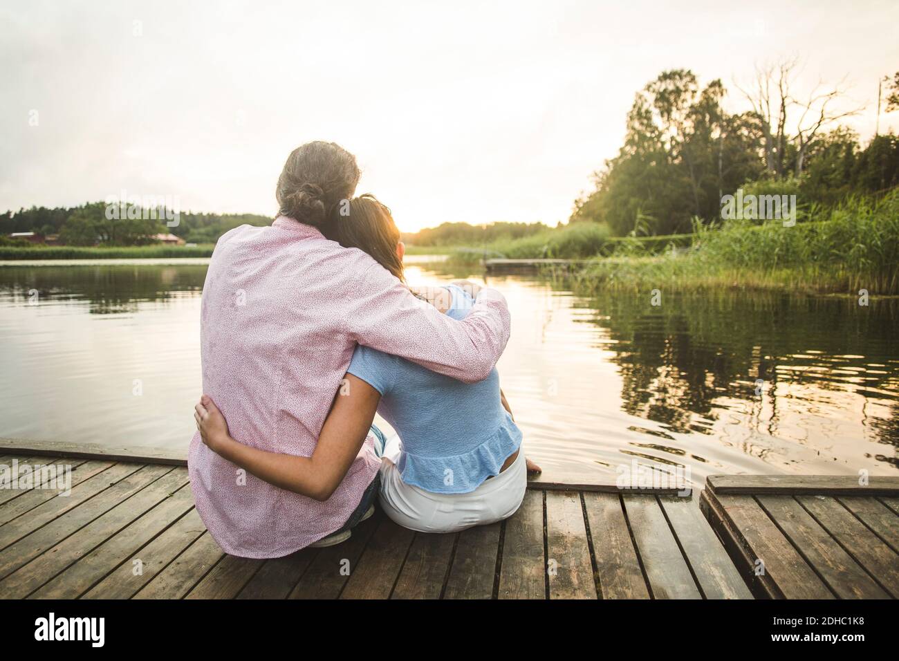 Vue arrière du couple assis avec les bras autour de la jetée au-dessus du lac au coucher du soleil Banque D'Images