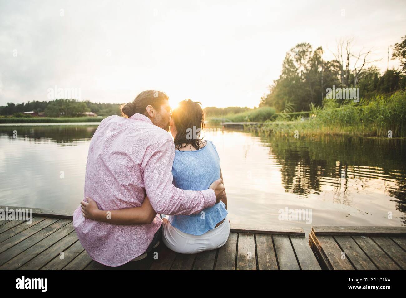 Vue arrière de l'ami embrassant petite amie assis avec les bras autour sur la jetée au-dessus du lac pendant le coucher du soleil Banque D'Images