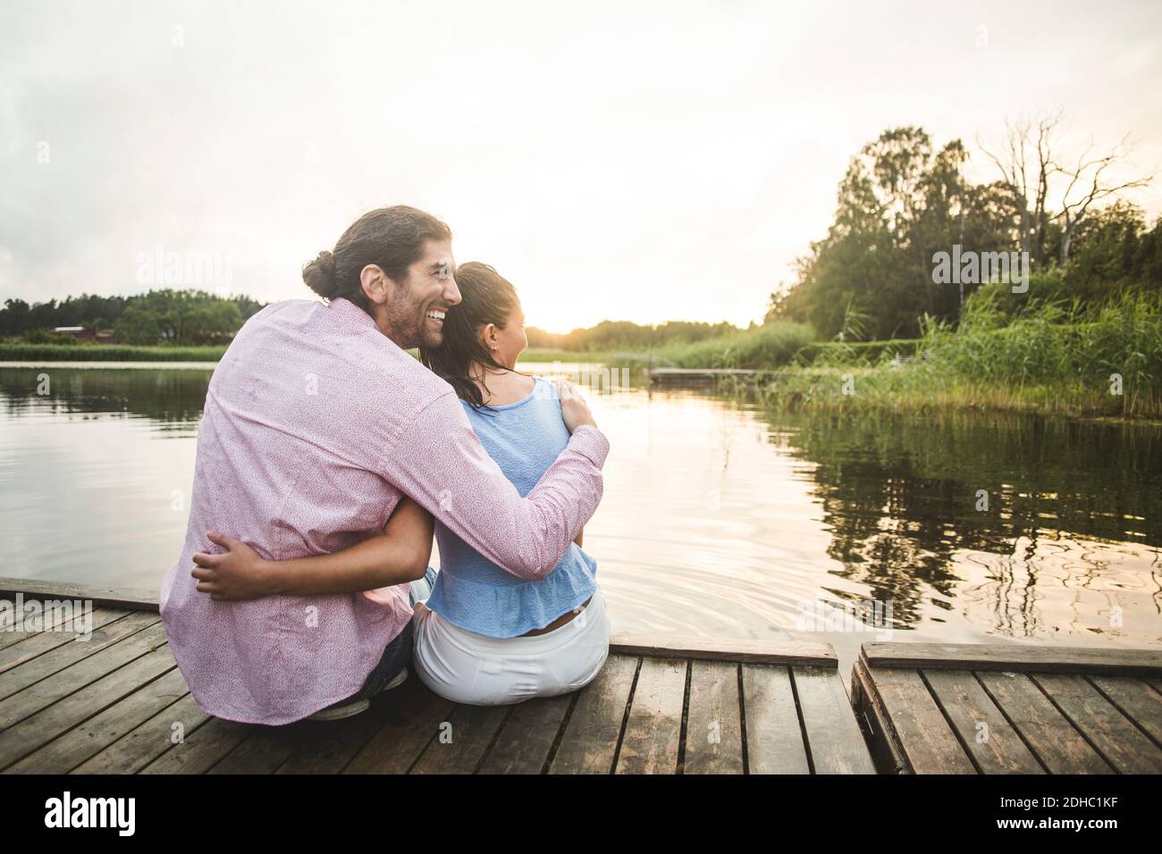 Vue arrière d'un couple souriant assis avec les bras jetée sur le lac au coucher du soleil Banque D'Images