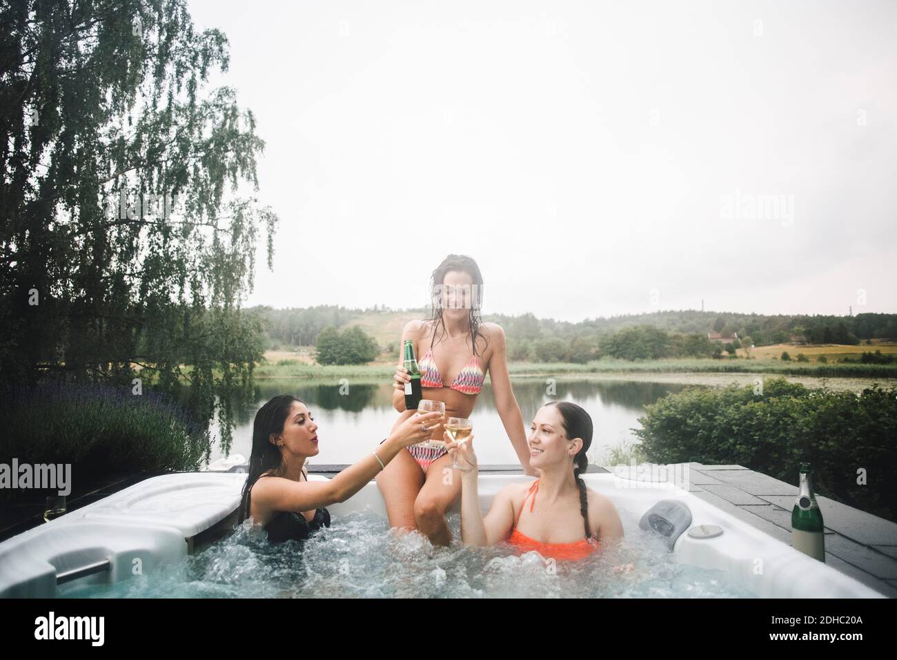 Des amies gaies toasting boissons dans le bain à remous contre clair ciel pendant une escapade d'un week-end Banque D'Images