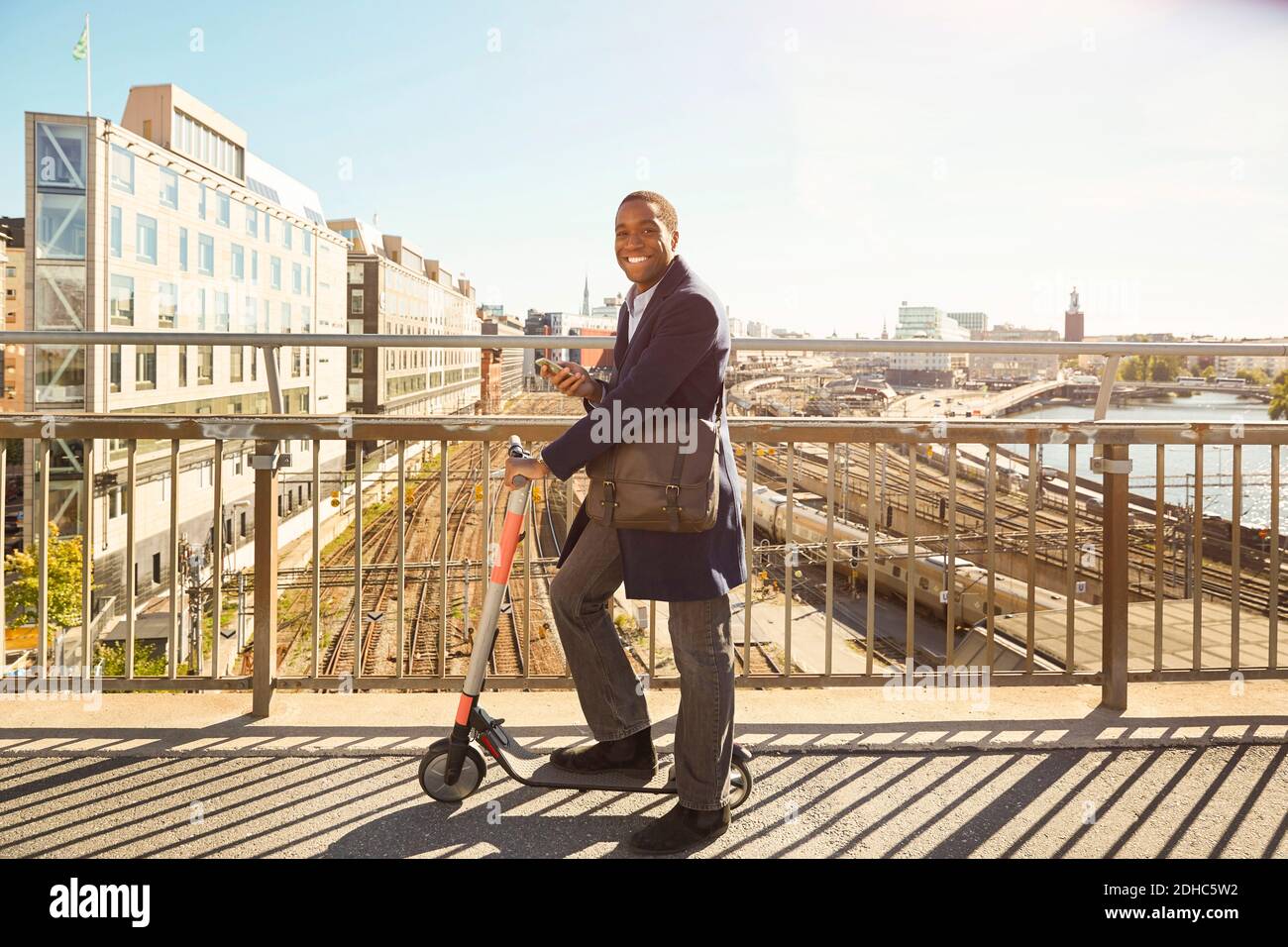 Vue latérale d'un jeune homme souriant debout avec un appareil électrique pousser le scooter sur le pont en ville Banque D'Images