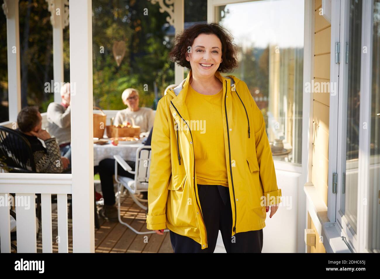 Portrait d'une femme mûre et gaie portant une veste jaune debout sur le porche avec des amis assis en arrière-plan Banque D'Images