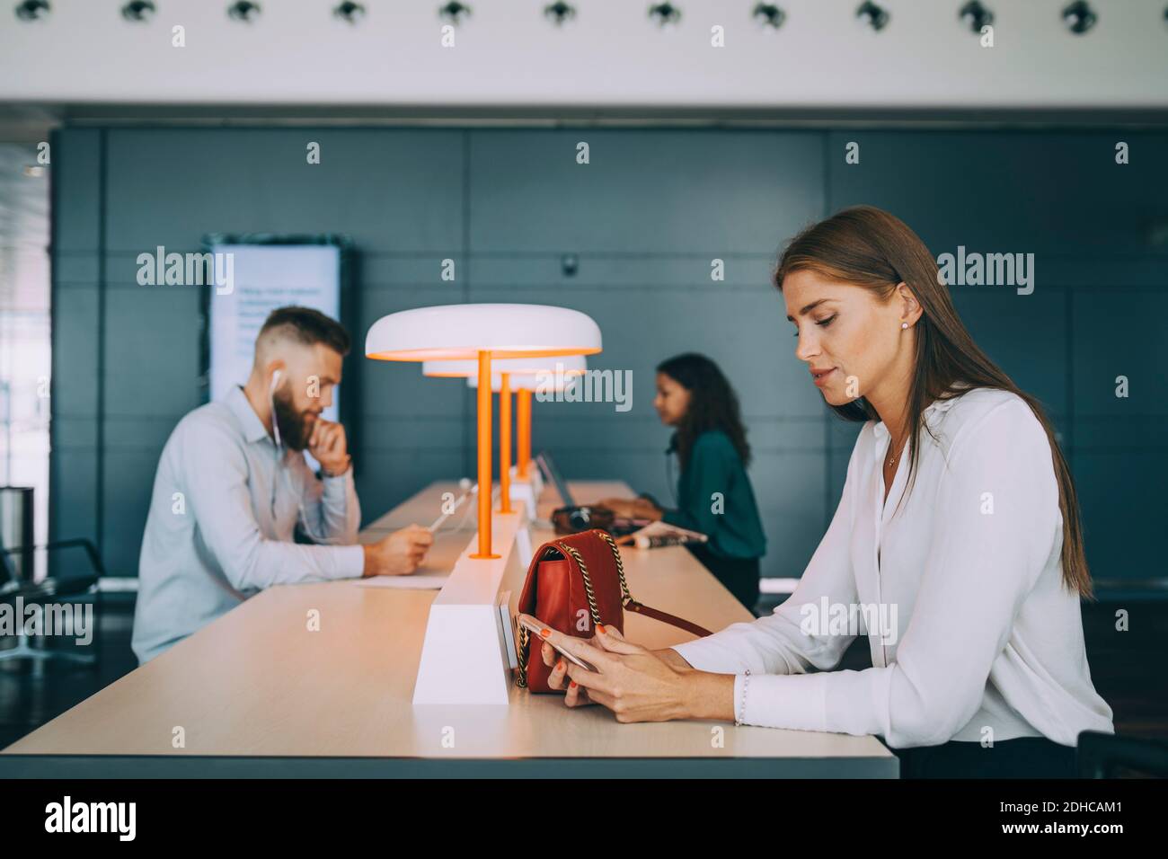 Jeune femme d'affaires utilisant un téléphone intelligent tout en étant assise avec des collègues multiethniques à la table d'attente au départ de l'aéroport a Banque D'Images