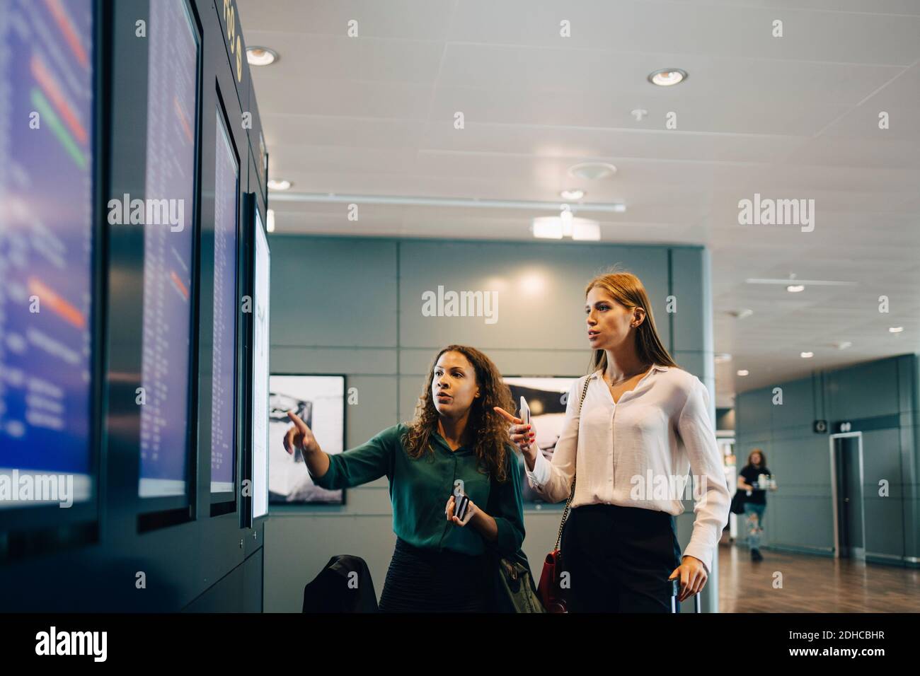 Des collègues féminins multiethniques examinent les horaires de vol sur le panneau de départ à l'arrivée à l'aéroport Banque D'Images