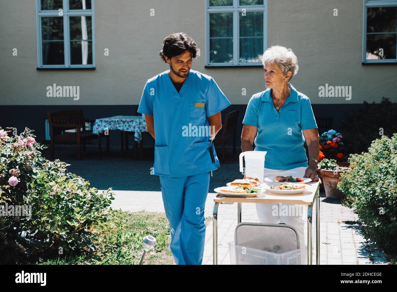 Une infirmière de sexe masculin marche avec une femme âgée poussant le chariot alimentaire contre maison de soins Banque D'Images