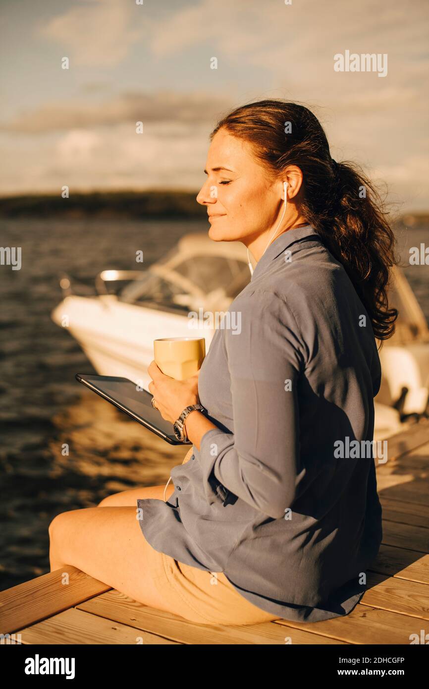 Femme mûre souriante prenant un café tout en utilisant une tablette numérique à patio contre le lac pendant les vacances Banque D'Images Femme mûre souriante prenant un café tout en utilisant une tablette numérique à patio contre le lac pendant les vacances Banque D'Images