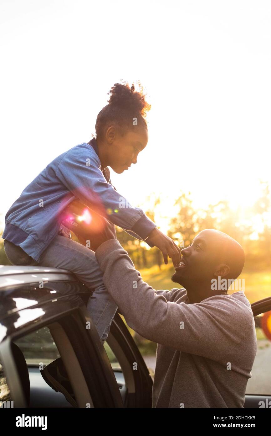 Homme souriant aidant la jeune fille à descendre du toit de voiture contre un ciel dégagé pendant le coucher du soleil au parc Banque D'Images