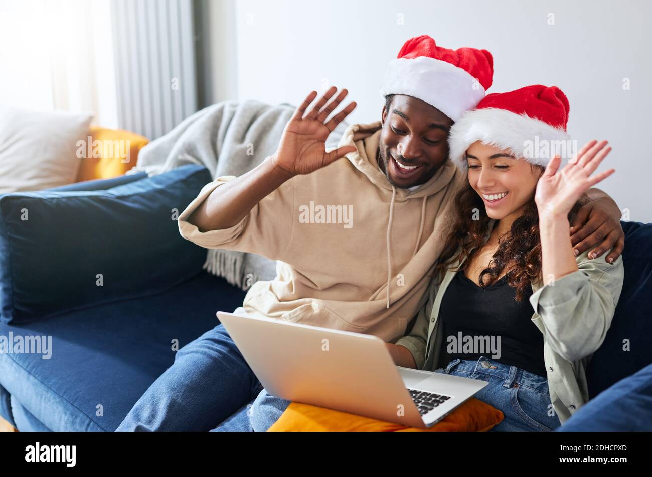 Un jeune couple ethnique mixte portant des chapeaux de père Noël se détende sur un canapé À la maison, passez des appels vidéo de Noël à la famille sur un ordinateur portable et amis Banque D'Images