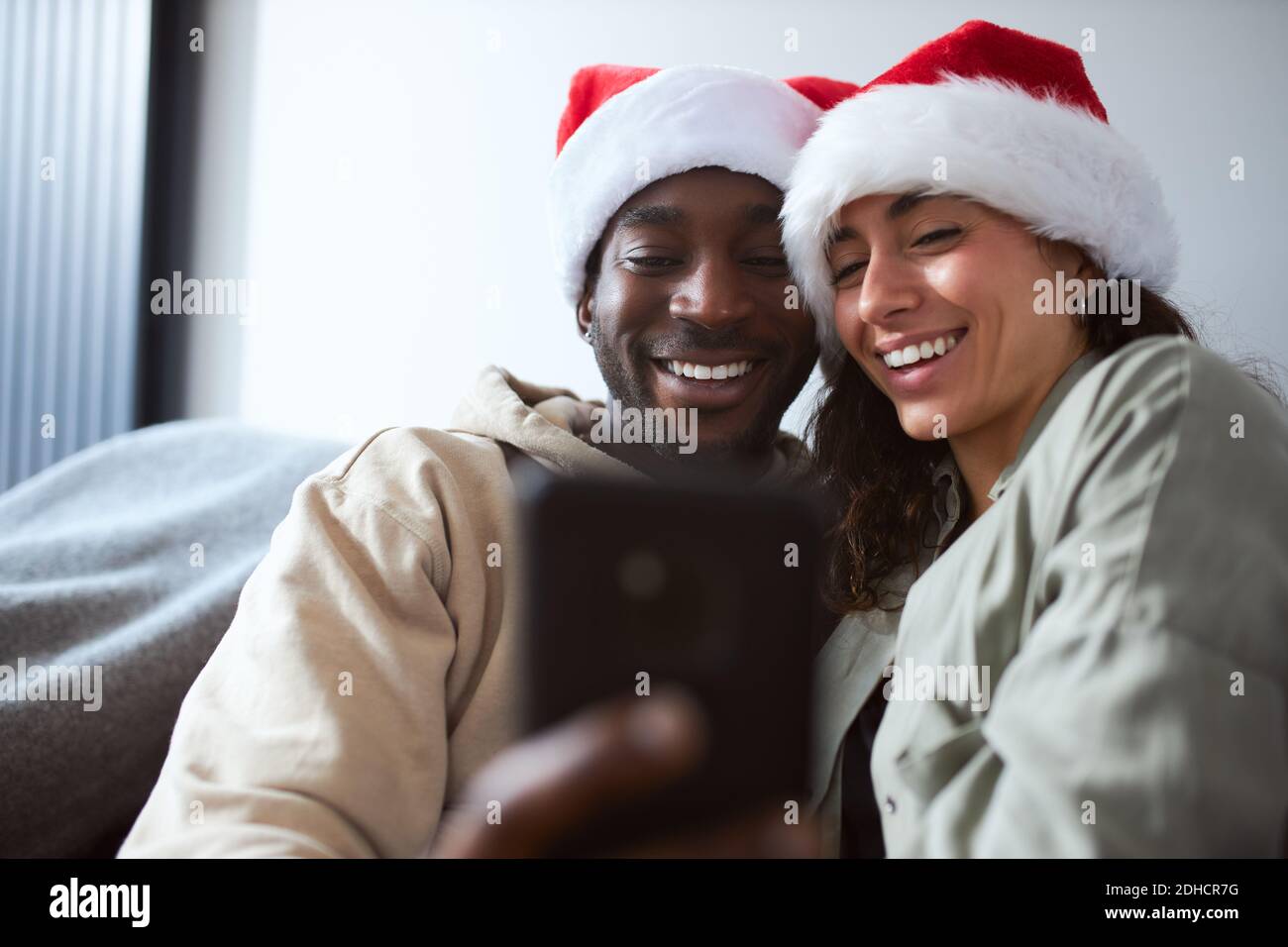 Jeune couple mixte d'ethnie se détendant sur le canapé à la maison portant chapeau de père noël posant pour le selfie de noël sur le téléphone mobile Banque D'Images