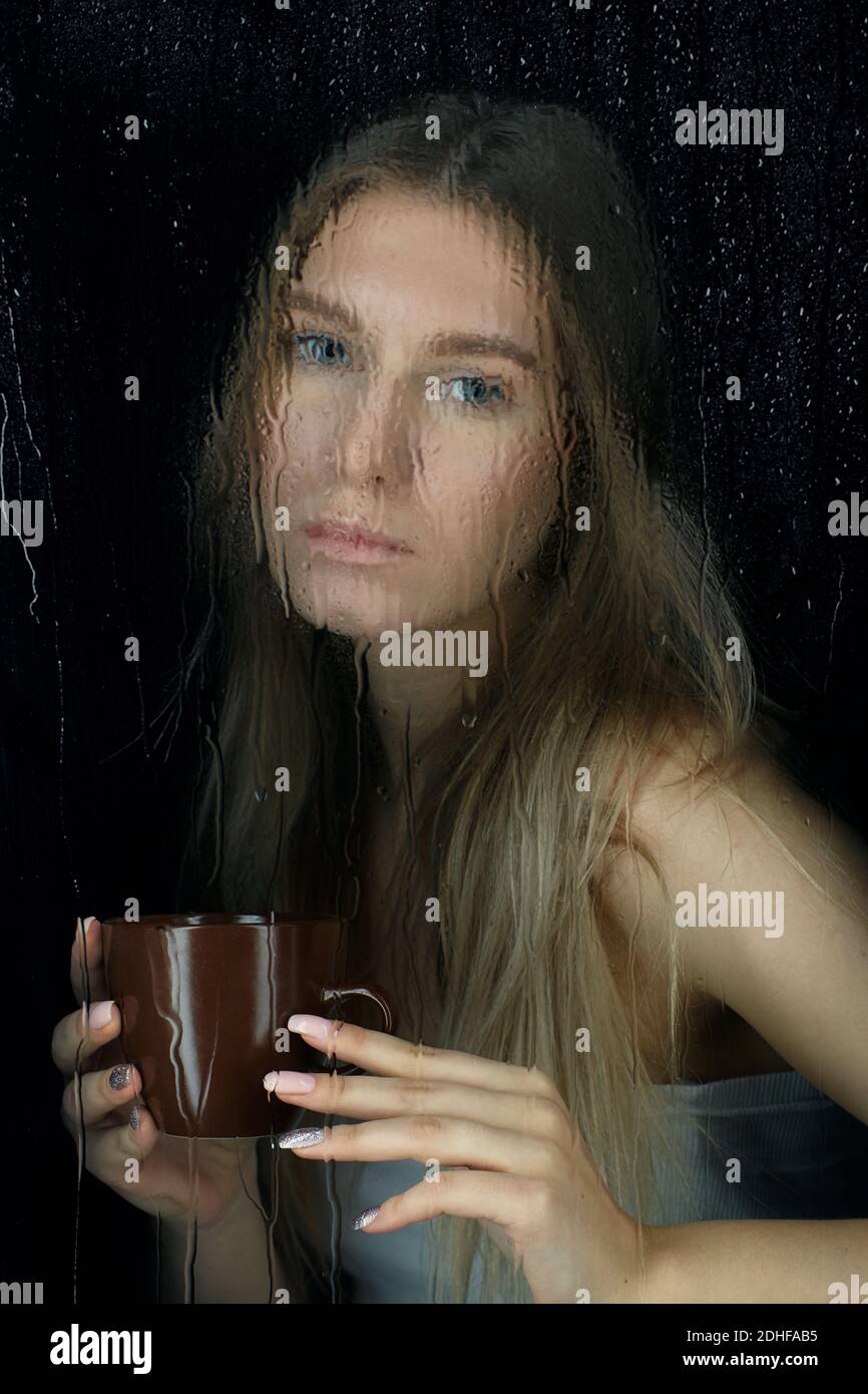 Verrouillage. Portrait d'une jeune femme blonde avec une tasse de café entre les mains. Femelle derrière la vitre avec gouttes de pluie. Banque D'Images