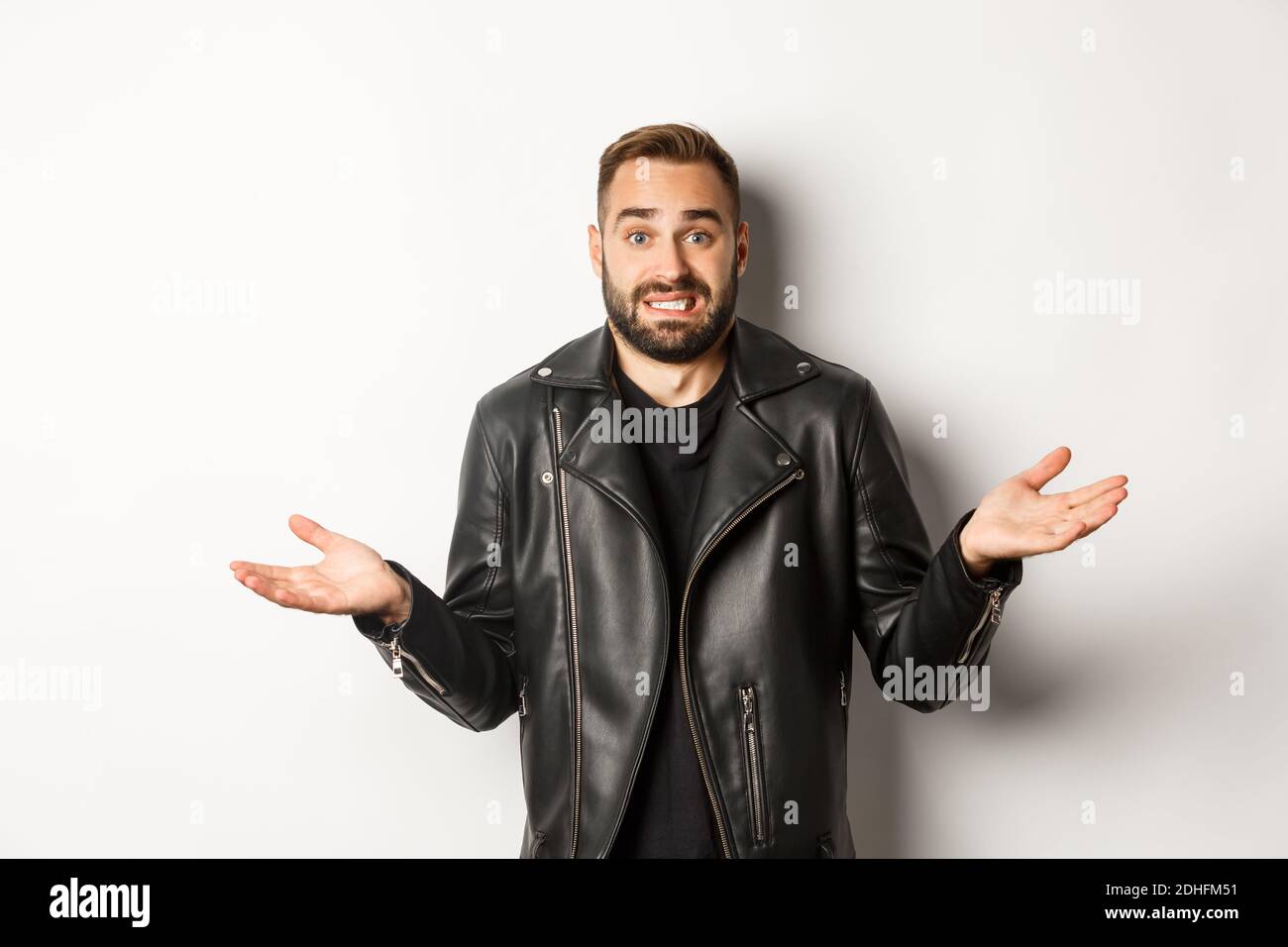 Homme barbu dans une veste en cuir noir demandant des conseils, en apparence sans indice et haussant, debout sur fond blanc Banque D'Images
