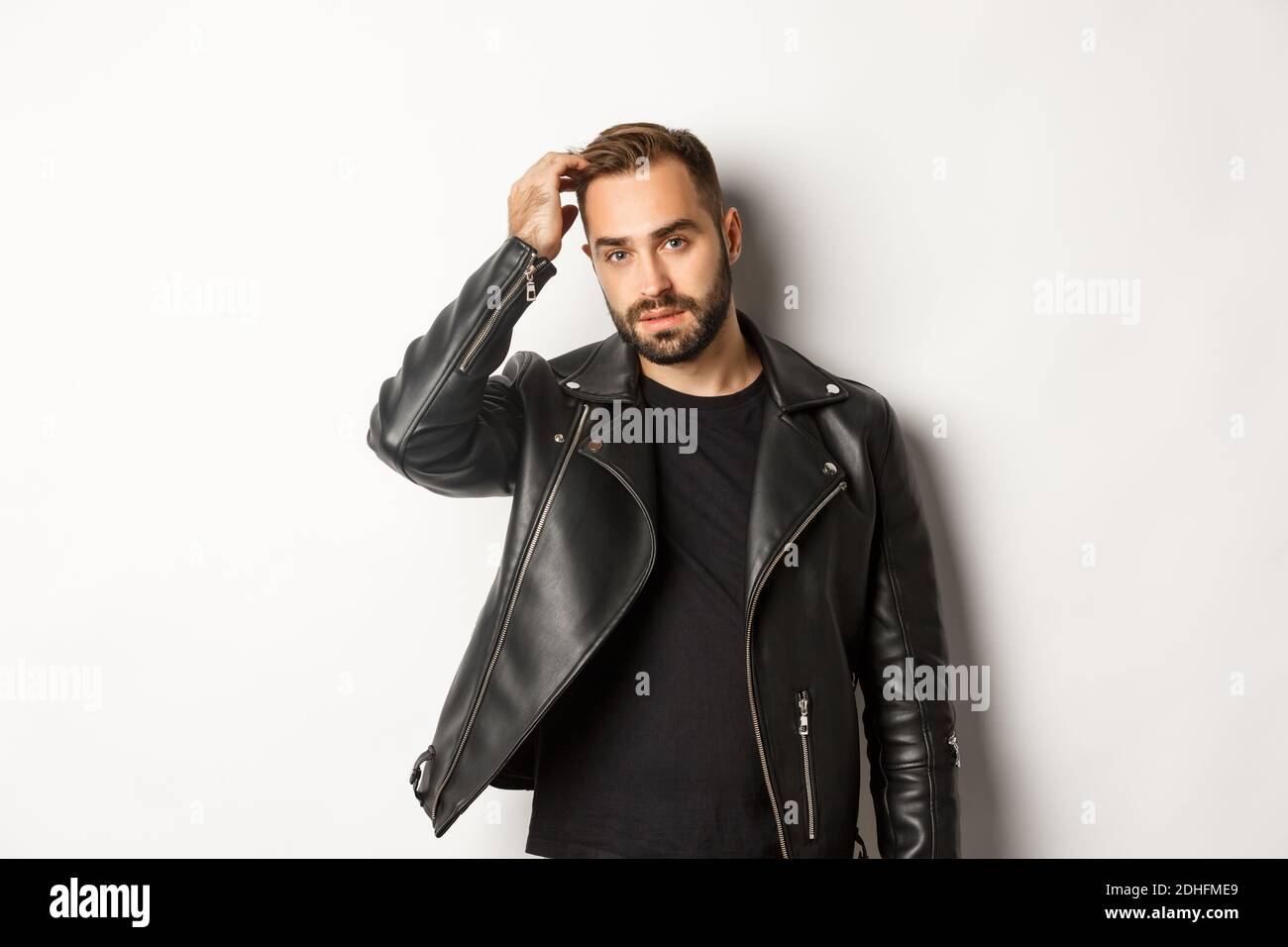 Beau macho homme en blouson de motard noir, touchant sa coupe de cheveux et regardant cool, fond blanc Banque D'Images