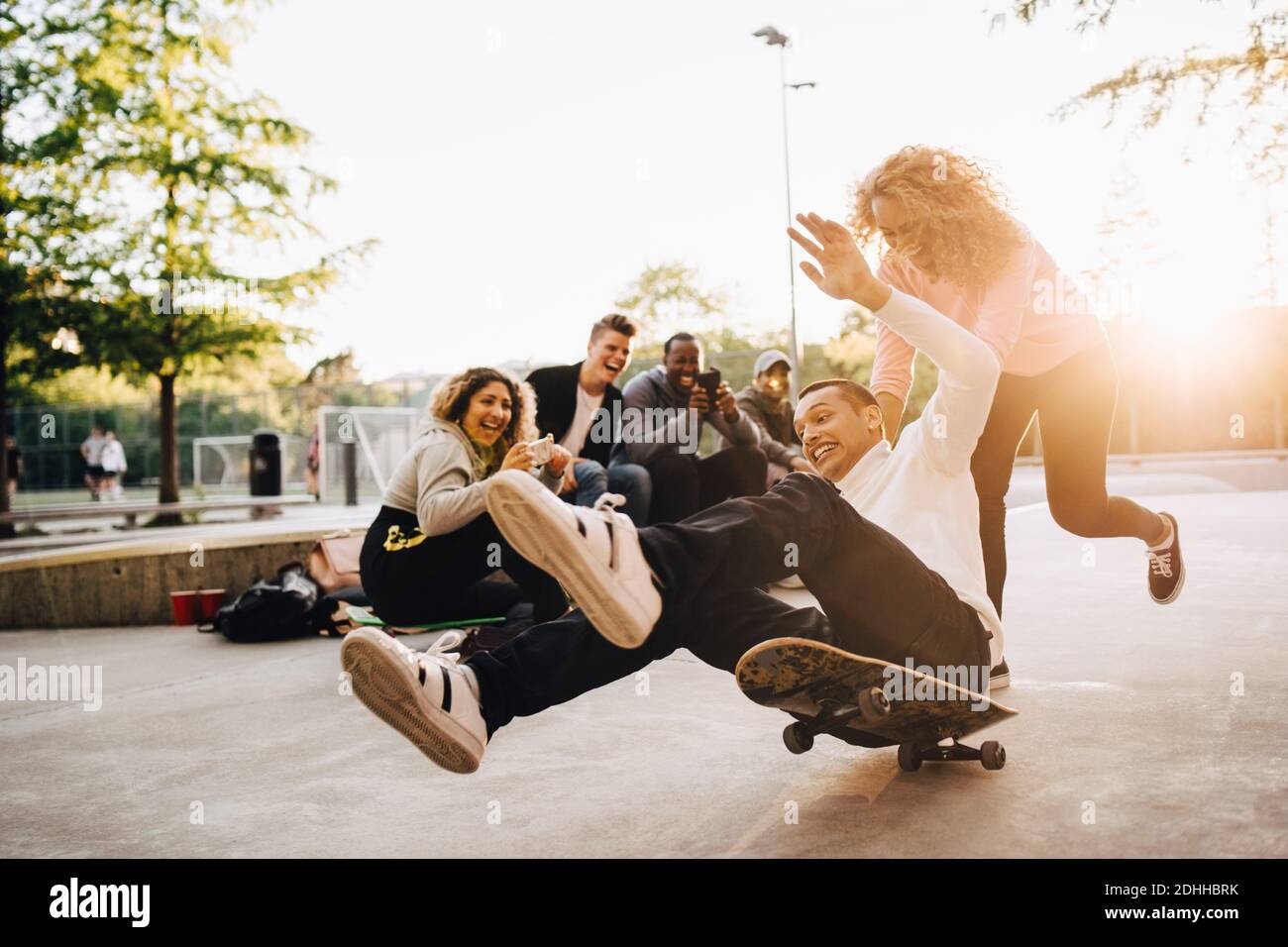Rire des amis photographier un homme tombant du skateboard pendant que la femme pousse lui au parc Banque D'Images