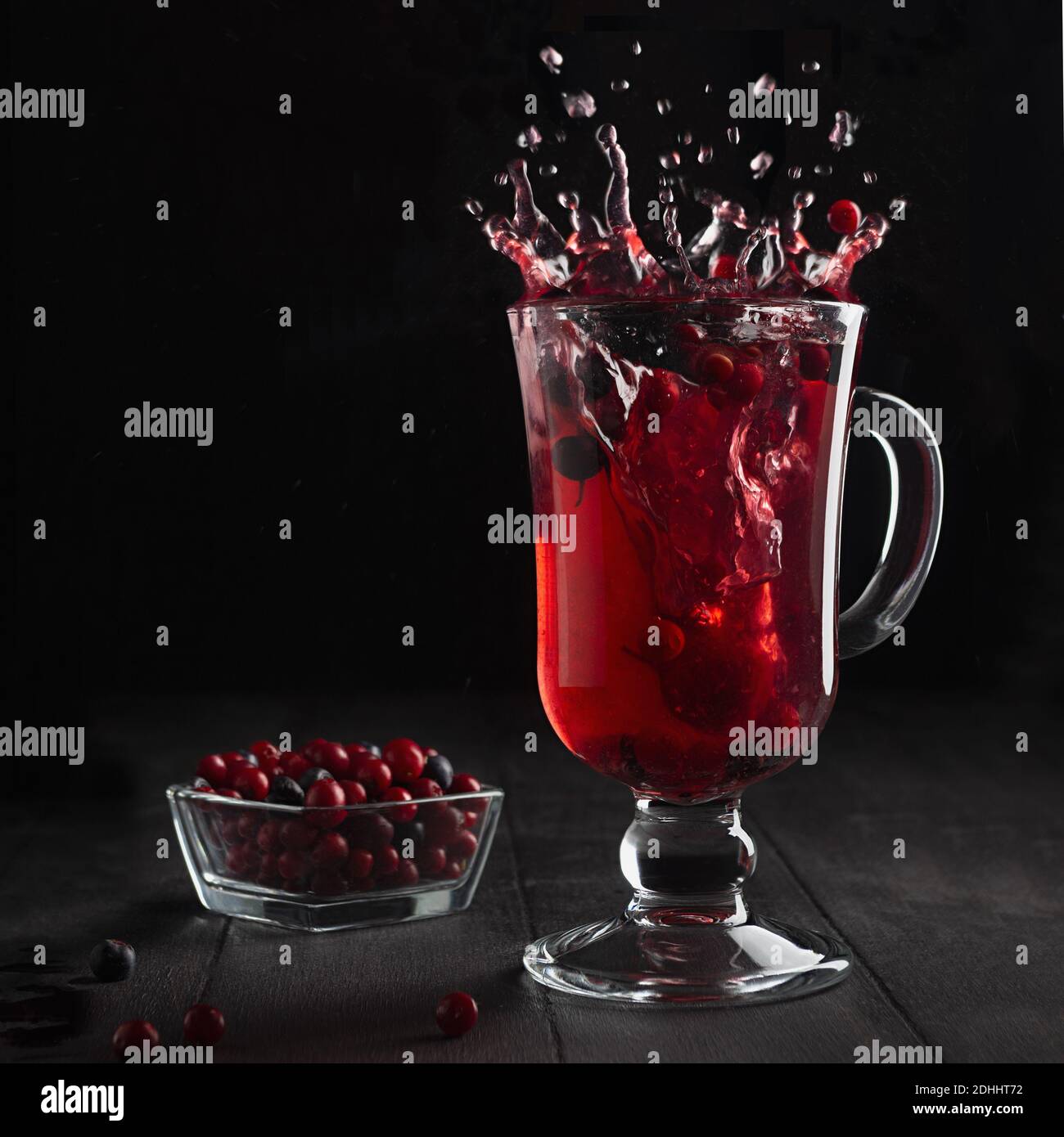 Thé aux baies rouges avec des éclaboussures élégantes et tourbillon dans verre de vin avec baie de cowberry mûre, myrtille sur table en bois noir dans intérieur noir foncé, carré. Banque D'Images