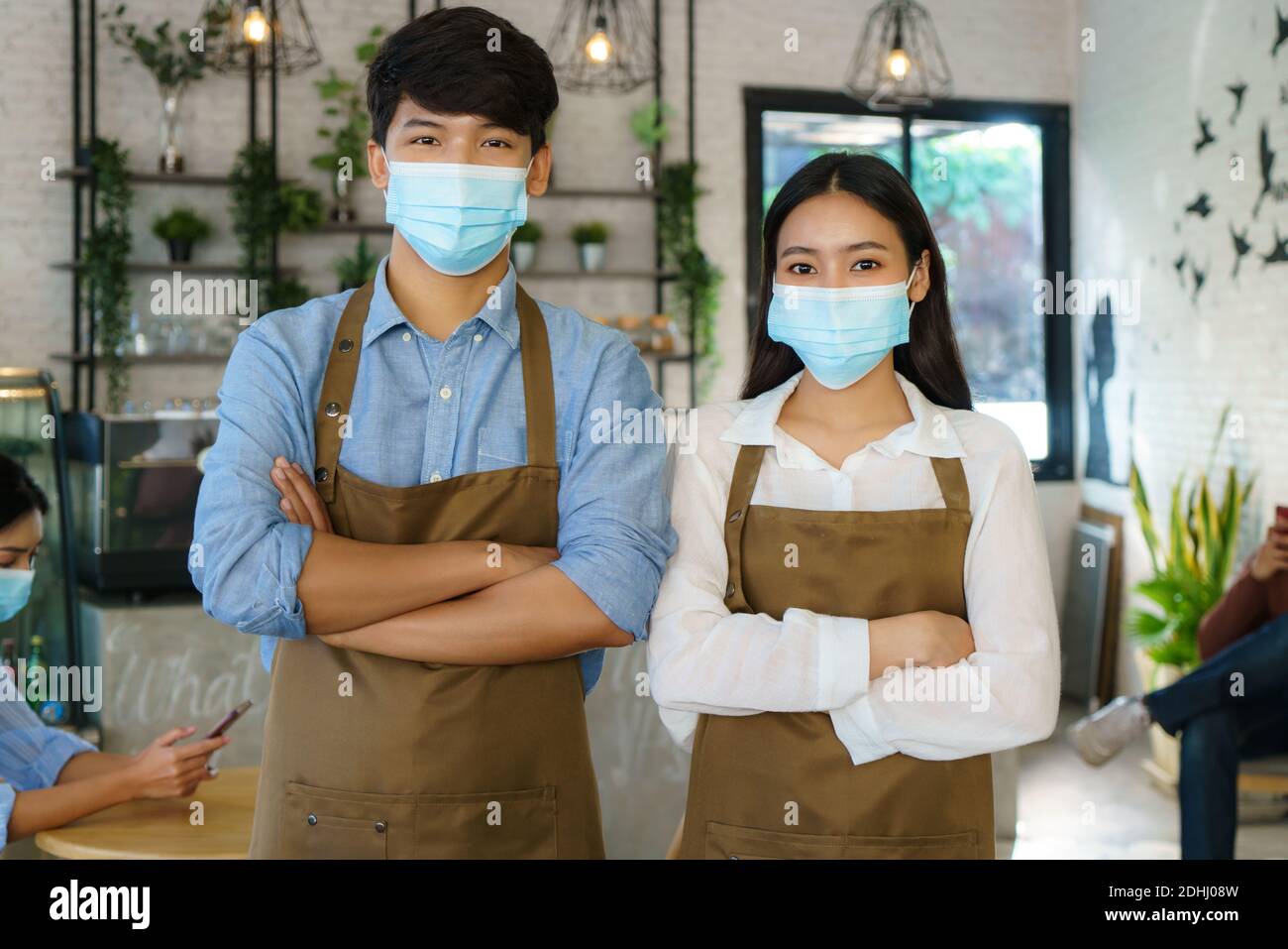 Couple de Barista asiatique en tablier et masque de visage regardant Appareil photo prêt à offrir un service de café au café moderne boutique Banque D'Images