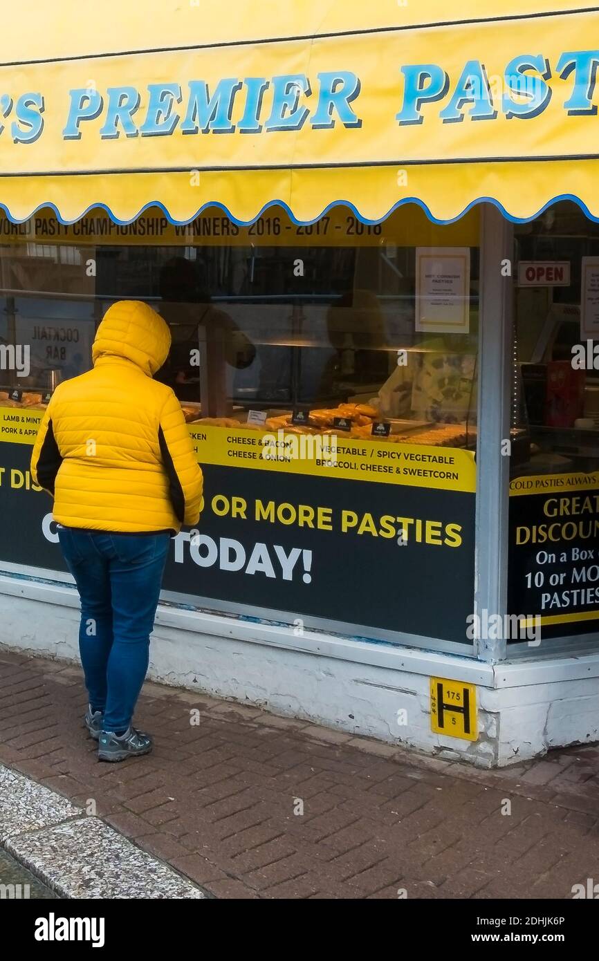 Une femme portant une veste jaune regardant dans la fenêtre d'un magasin de pasties de Cornouailles avec un panneau jaune à Newquay en Cornouailles. Banque D'Images