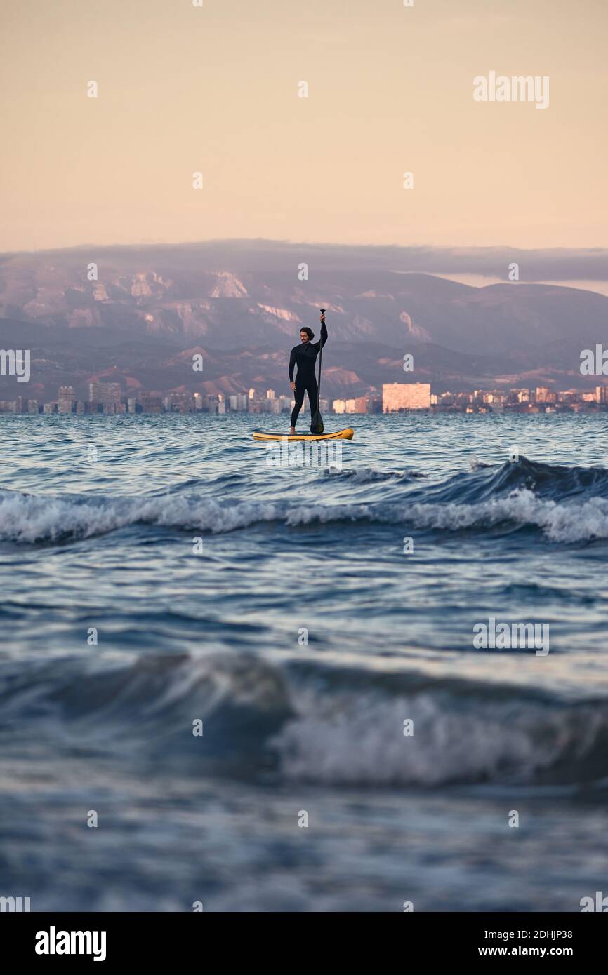 Surfeur mâle en combinaison avec aviron à paddle-board en mer eau sur fond de montagnes au coucher du soleil Banque D'Images